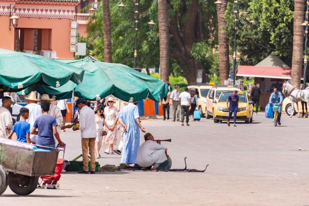 Busy sunlit square in Marrakech with yellow taxis, palm trees and street performers