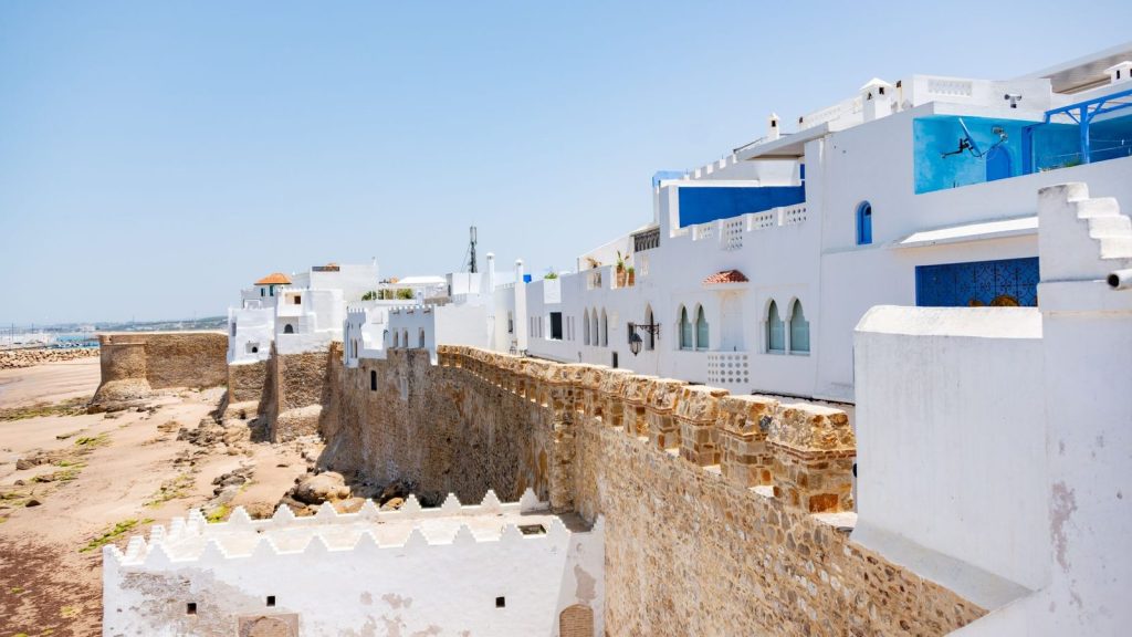 A view of a whitewashed medina with an ancient seaside fortress under a clear blue sky in Morocco.