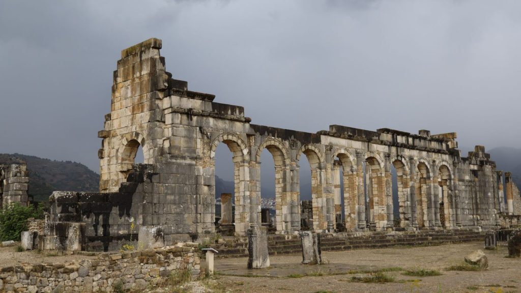 Ancient Roman ruins of Volubilis in Morocco, showcasing stone arches and dramatic cloudy sky.