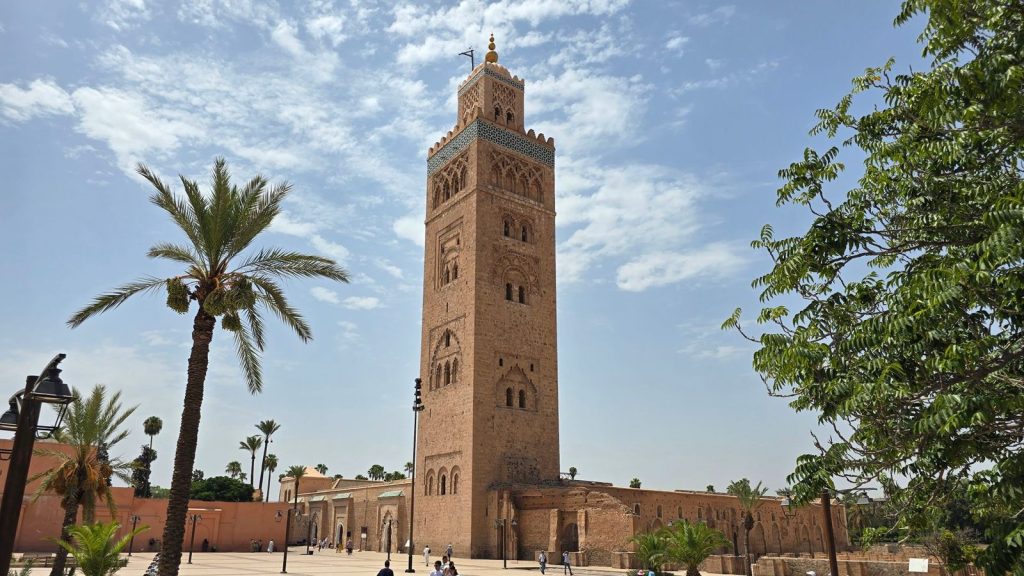 The Koutoubia Mosque minaret in Marrakech under a blue sky, with palm trees in the square below
