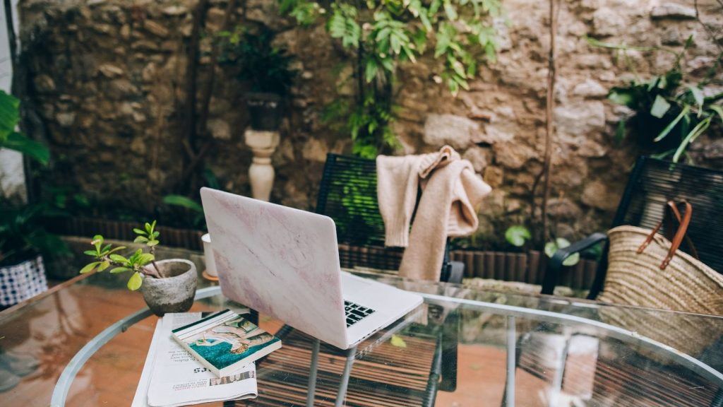 Open laptop on a table in a shaded stone courtyard with plants and a straw basket